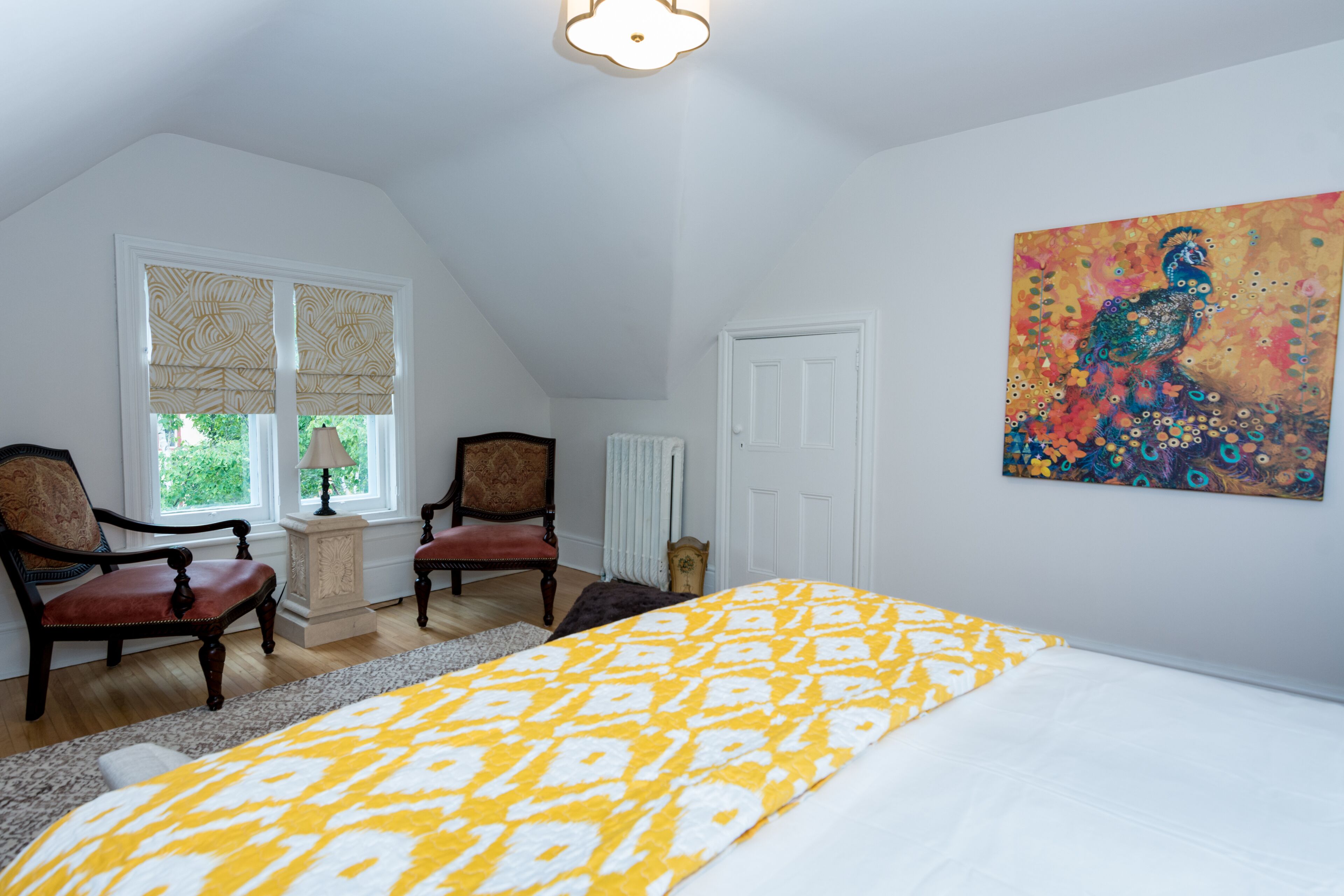 A bedroom with a bed, two chairs, a radiator, a window with blinds, and a painting on the wall.