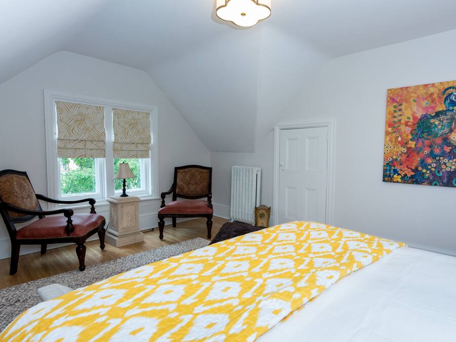 A bedroom with a bed, two chairs, a radiator, a window with blinds, and a painting on the wall.