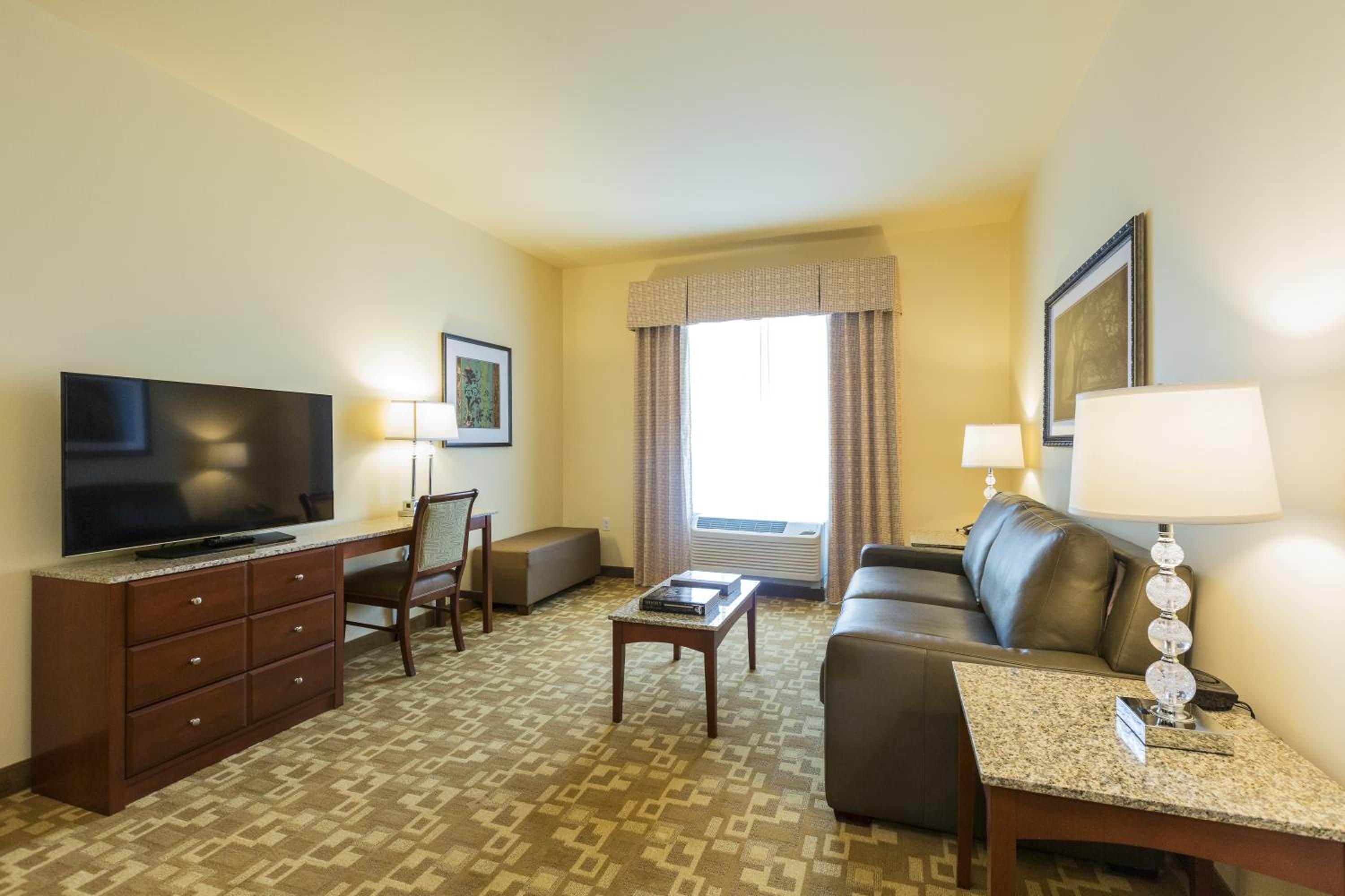 Une chambre d’hôtel équipée d’un téléviseur à écran plat, d’un canapé, d’une table basse, d’un bureau avec une chaise et d’une fenêtre avec des rideaux.