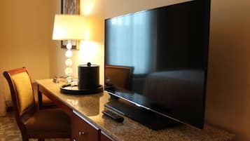 A hotel room with a large flat-screen TV on a granite countertop, a lamp, a chair, and a bed.