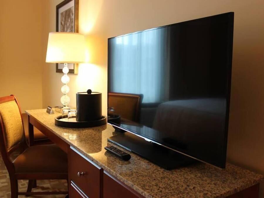 A hotel room with a large flat-screen TV on a granite countertop, a lamp, a chair, and a bed.