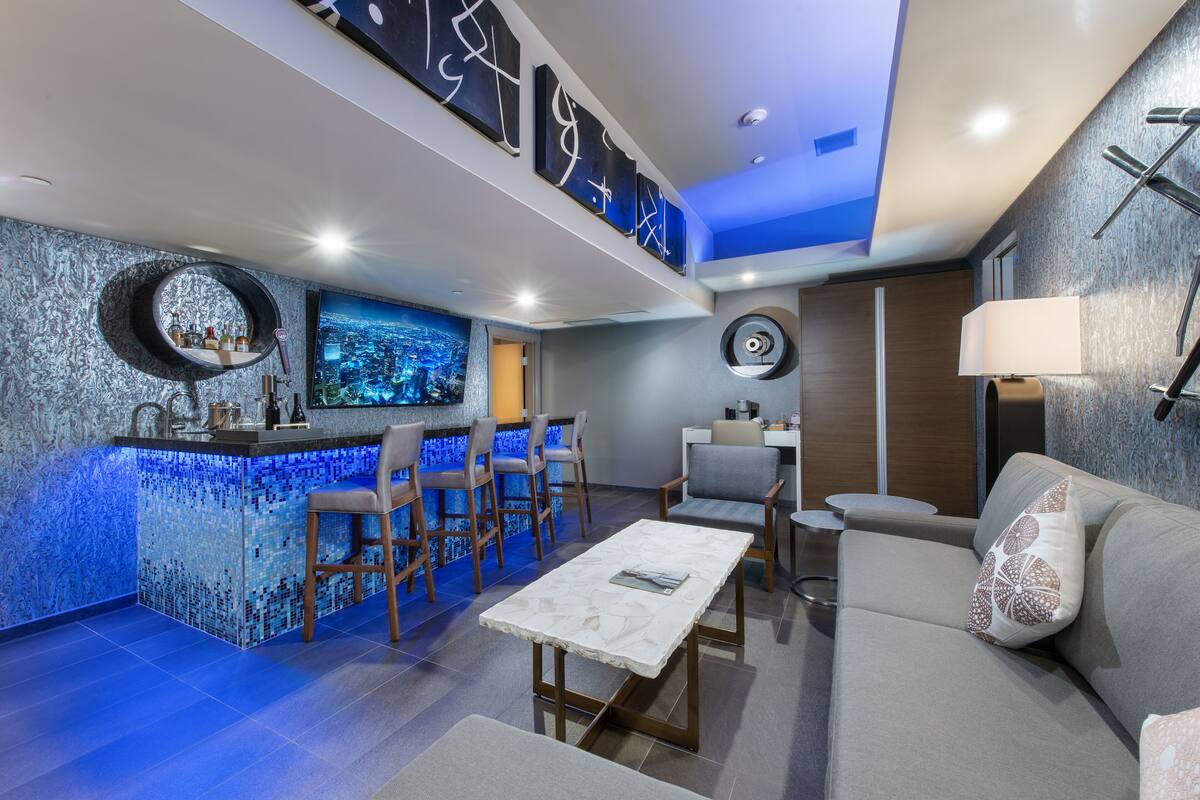 A modern bar area with a blue-lit counter, seating with bar stools, and a large screen displaying a cityscape.