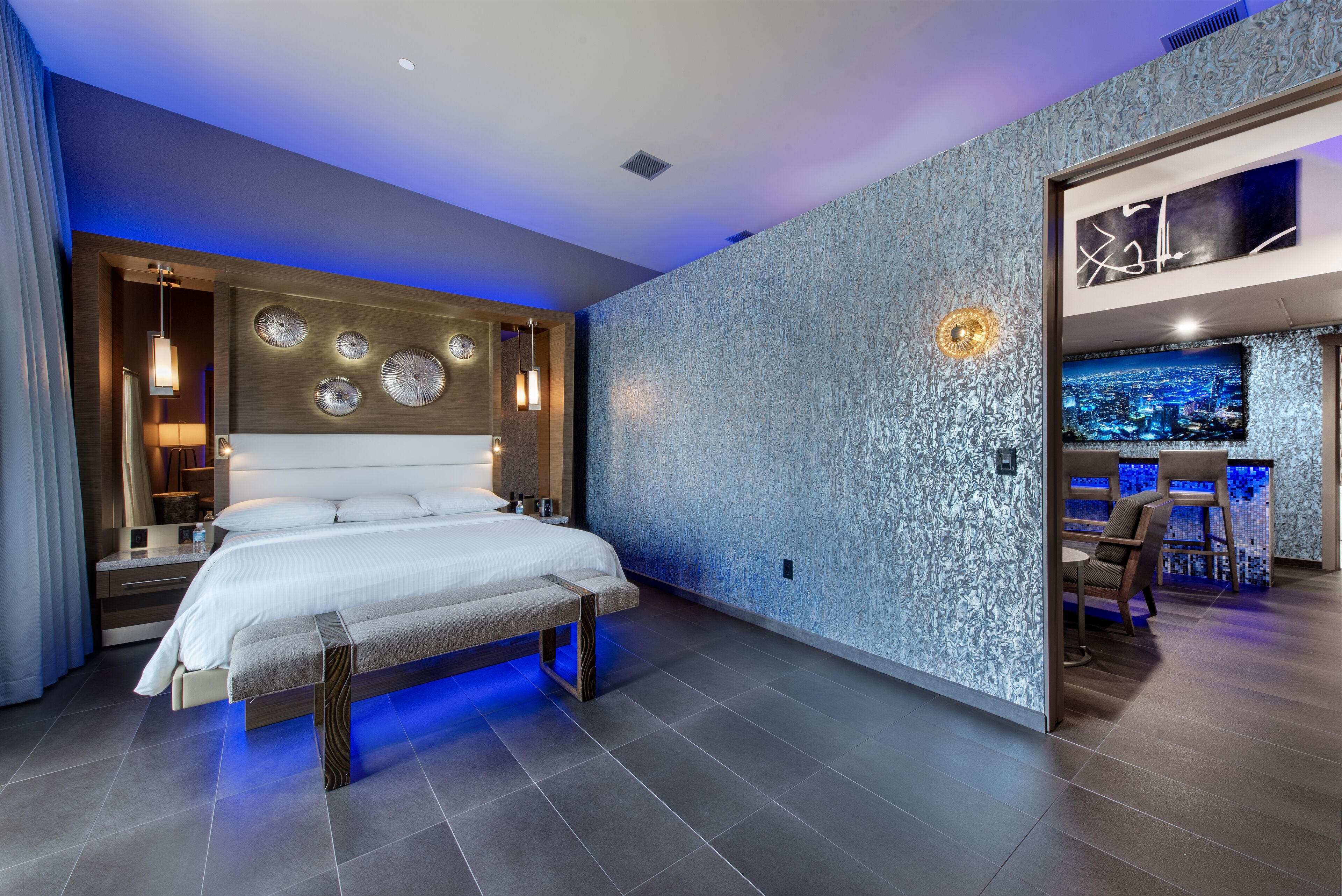 Una habitación de hotel moderna con una cama grande, una zona de estar con televisor y una pared con textura particular.
