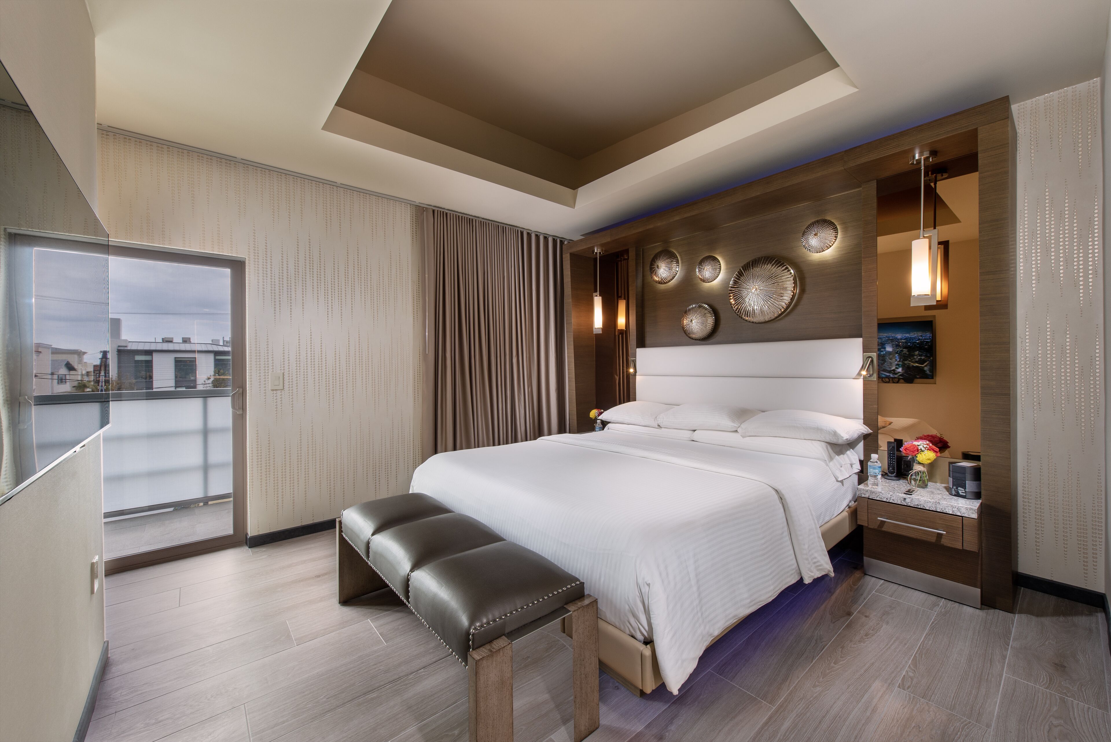 Habitación de hotel moderna con una cama grande, un banco, una mesita de noche y vistas a la ciudad.