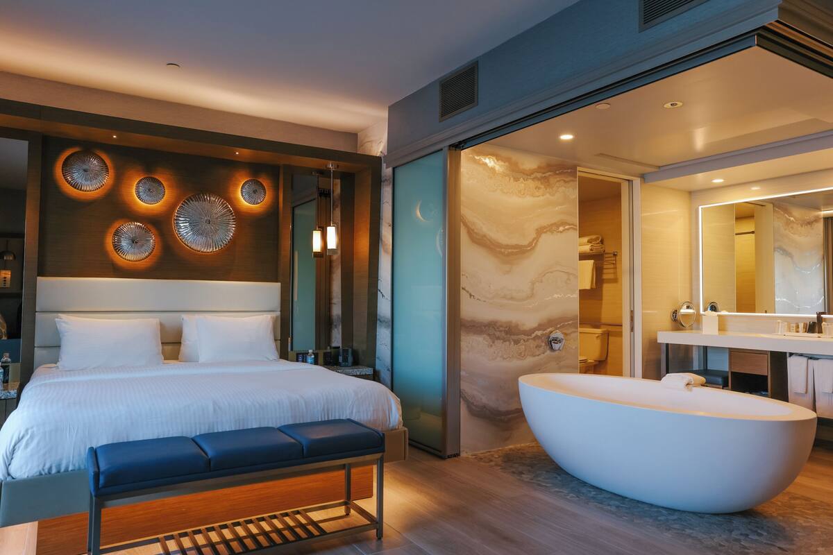A modern hotel room with a large bed, a built-in bench, a freestanding bathtub, and a wall with circular light fixtures.