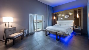 A modern hotel room with a large bed, a chair, a lamp, and a view of the outside through a glass door. - Shade Hotel (Manhattan Beach)