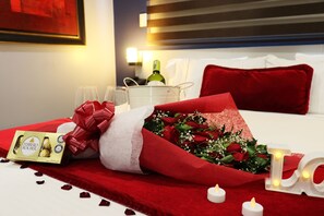 A hotel room with a bed, a bouquet of red roses, a bottle of champagne, and a gift box.