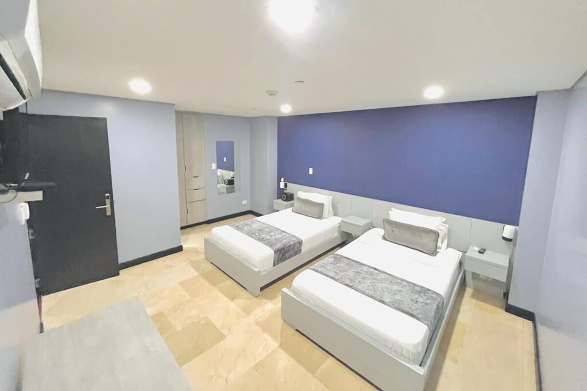 A modern hotel room with two beds, a dark door, a small bathroom, and a large window.