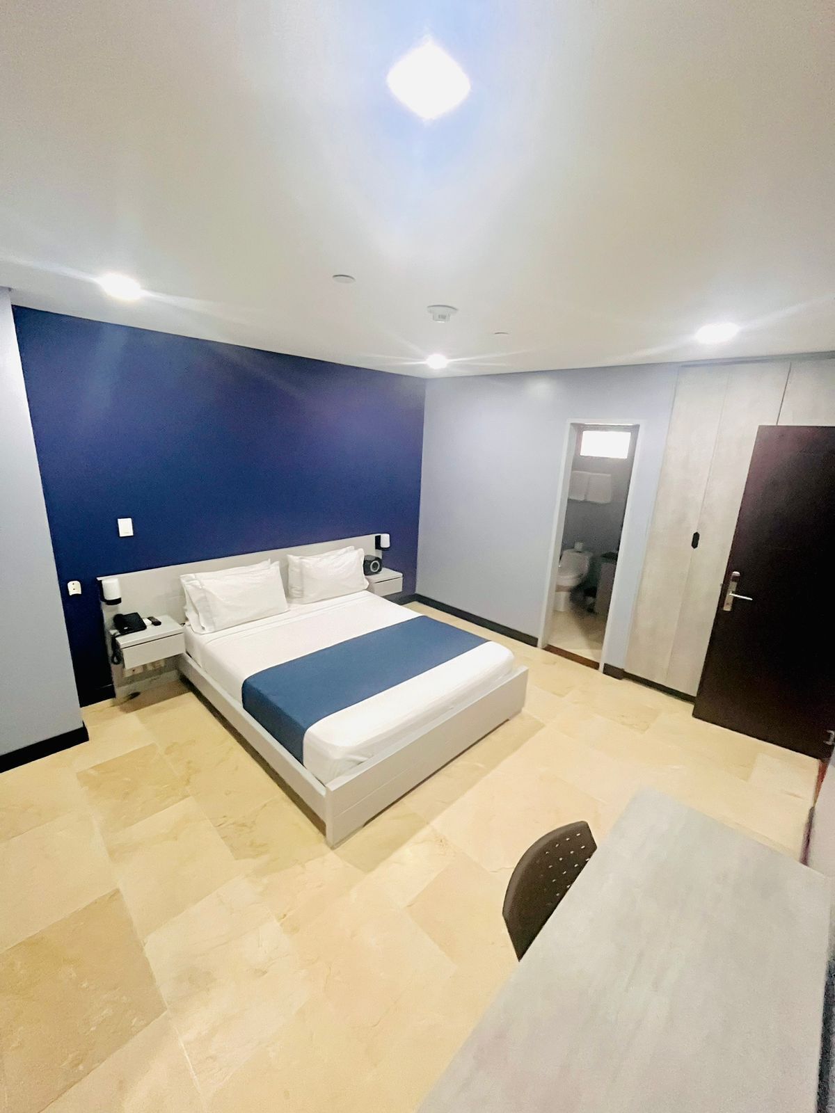 A modern hotel room with a large bed, a bedside table, a desk, and a wardrobe.