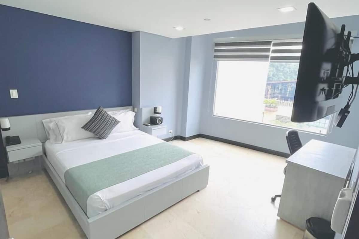 A modern hotel room with a large bed, a desk, a TV, and a window with blinds.