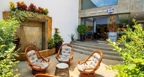 A hotel entrance with a stone facade, a signboard, and a seating area with wicker chairs and a table.