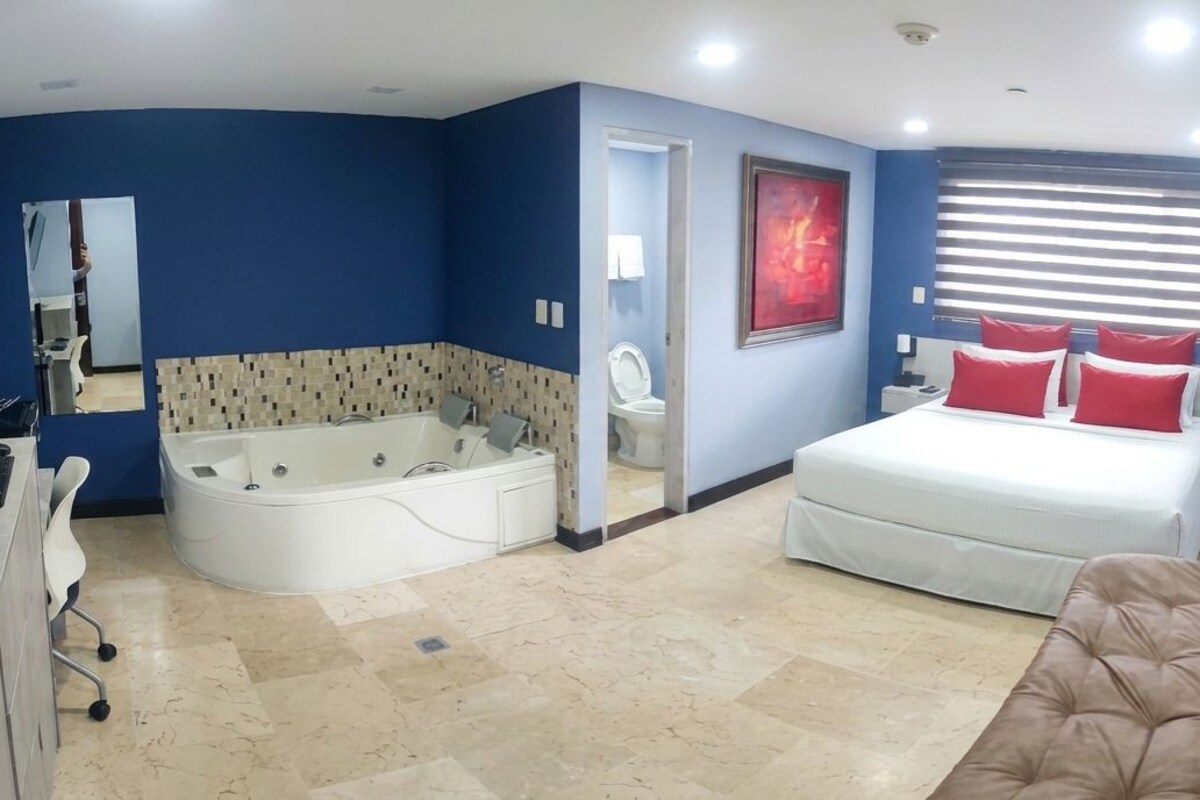 A bedroom with a bed, a sitting area, a desk, a bathtub, and a window with blinds.