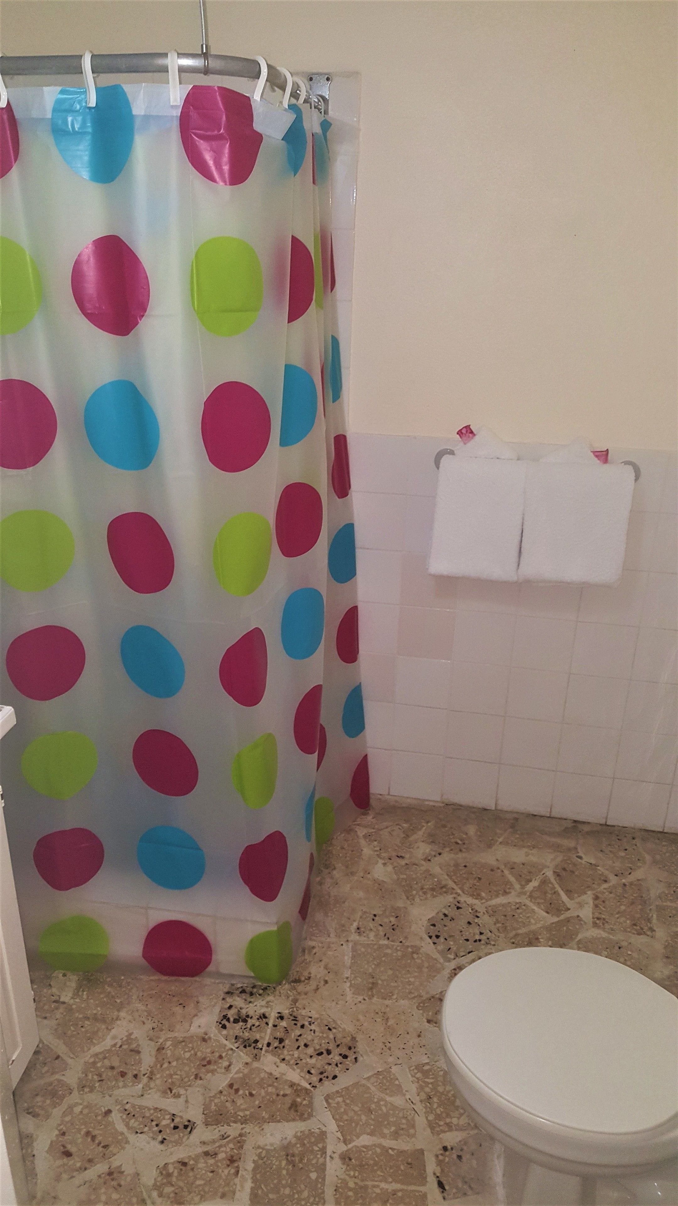 Un baño con cortina de ducha con lunares grandes en varios colores, pared de azulejos blancos, inodoro blanco y una toalla pequeña blanca colgada en la pared.