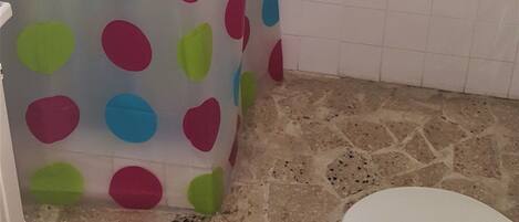 Un baño con cortina de ducha con lunares grandes en varios colores, pared de azulejos blancos, inodoro blanco y una toalla pequeña blanca colgada en la pared.