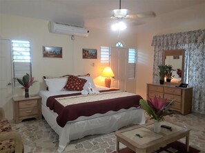 A bedroom with a bed, bedside tables, a nightstand, a dresser, a mirror, a ceiling fan, and a window with blinds. - Paradise Inn (Port Antonio)