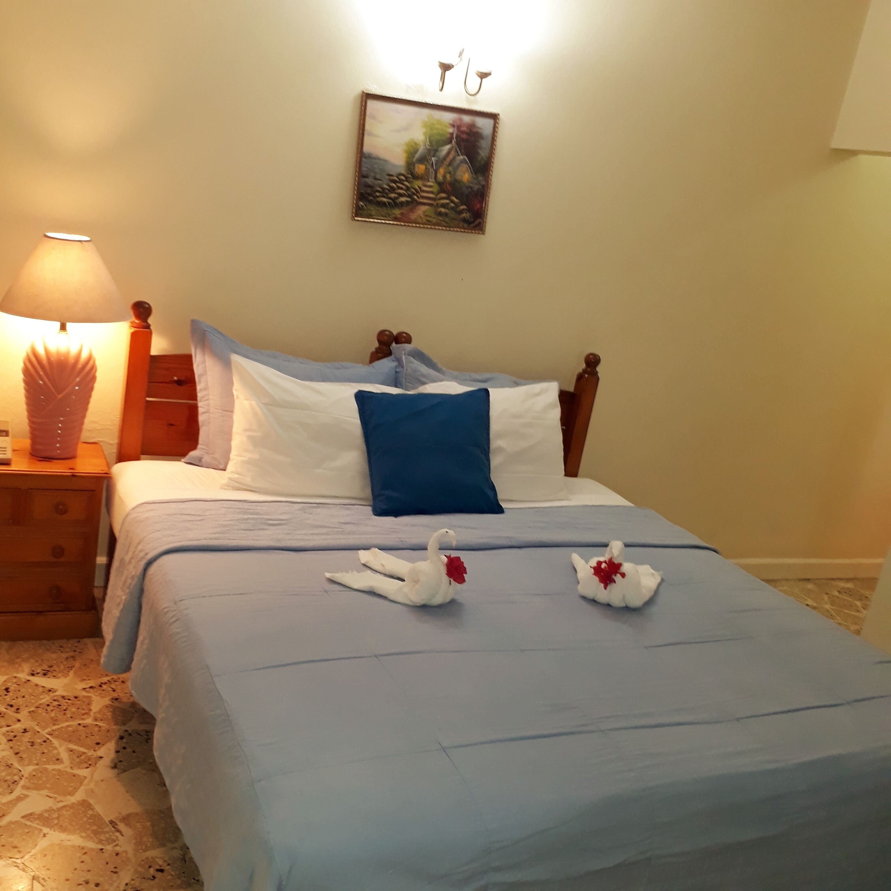 Una cama bien hecha con una colcha azul y almohadas blancas, dos almohadas con forma de cisne, un cabecero de madera, una mesita de noche con una lámpara y un cuadro en la pared.