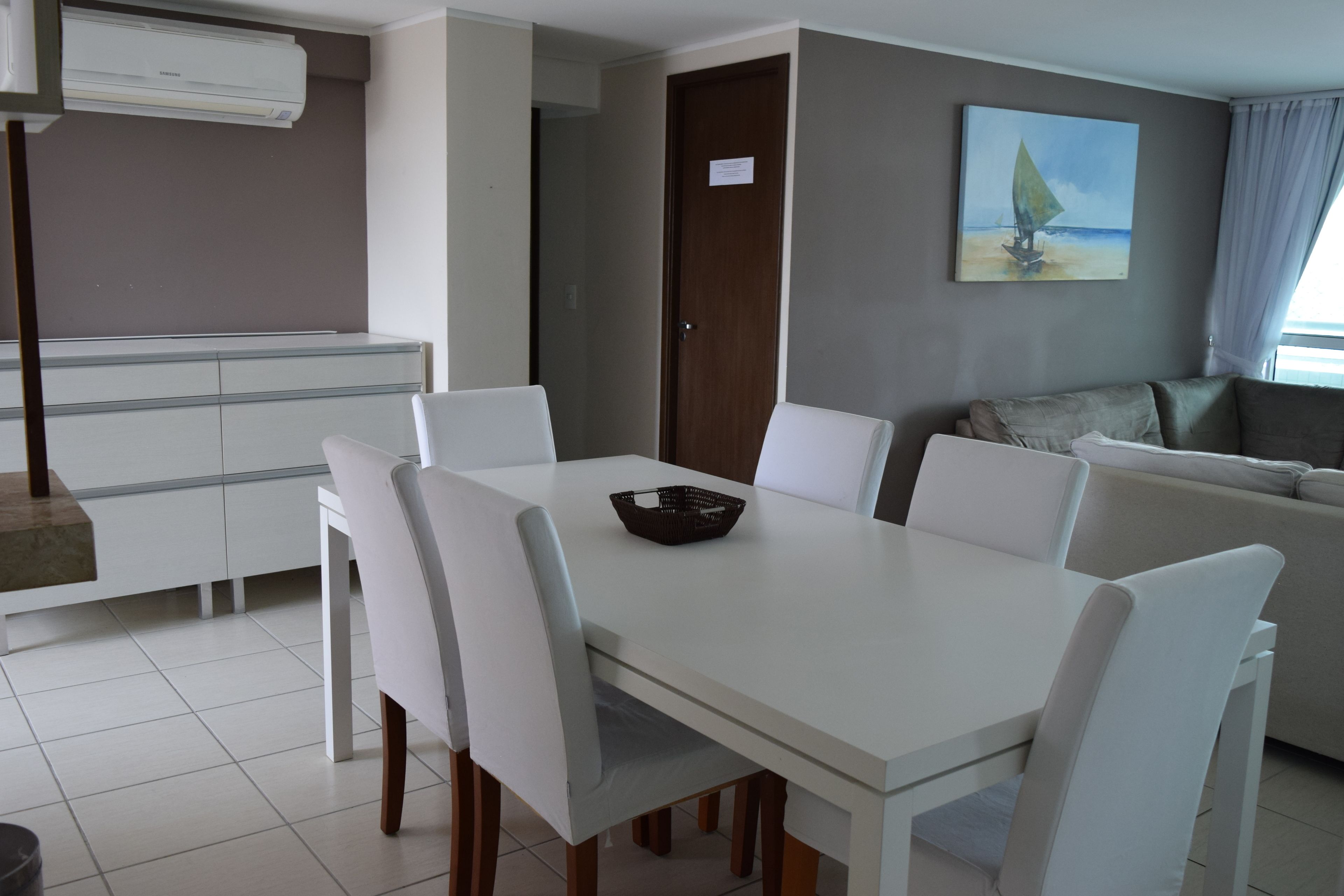 Uma sala de estar moderna com uma mesa de jantar branca, cadeiras brancas, um sofá cinzento e um quadro de um veleiro na parede.