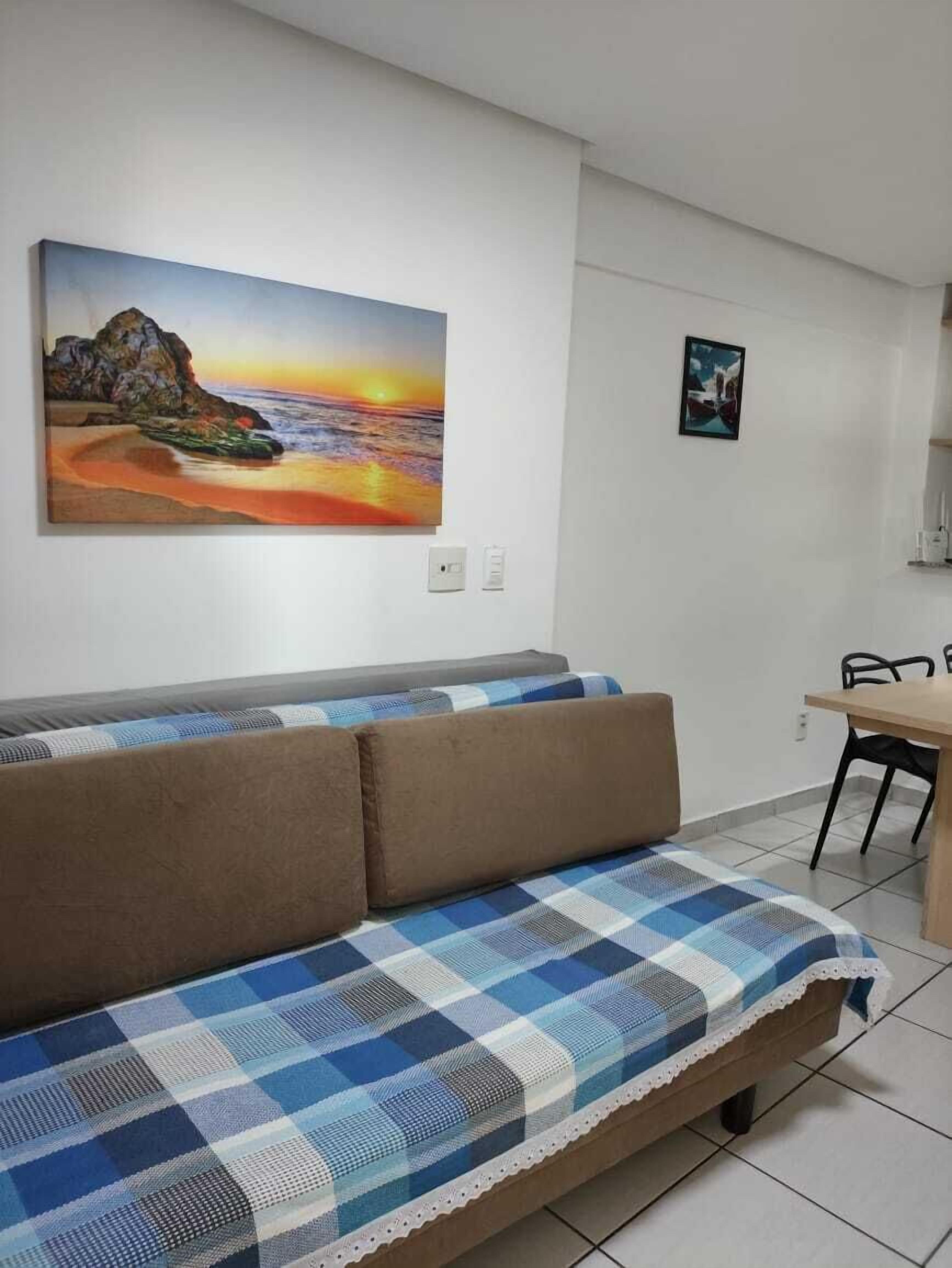 A living room with a sofa, a checkered blanket, a painting of a sunset, and a dining area with chairs.
