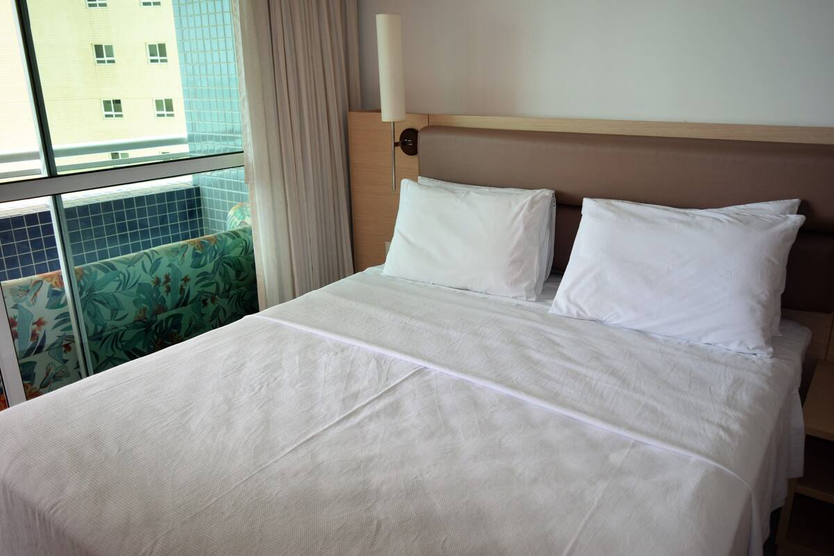A hotel room with a large bed, a nightstand, a wall-mounted lamp, and a window with curtains.