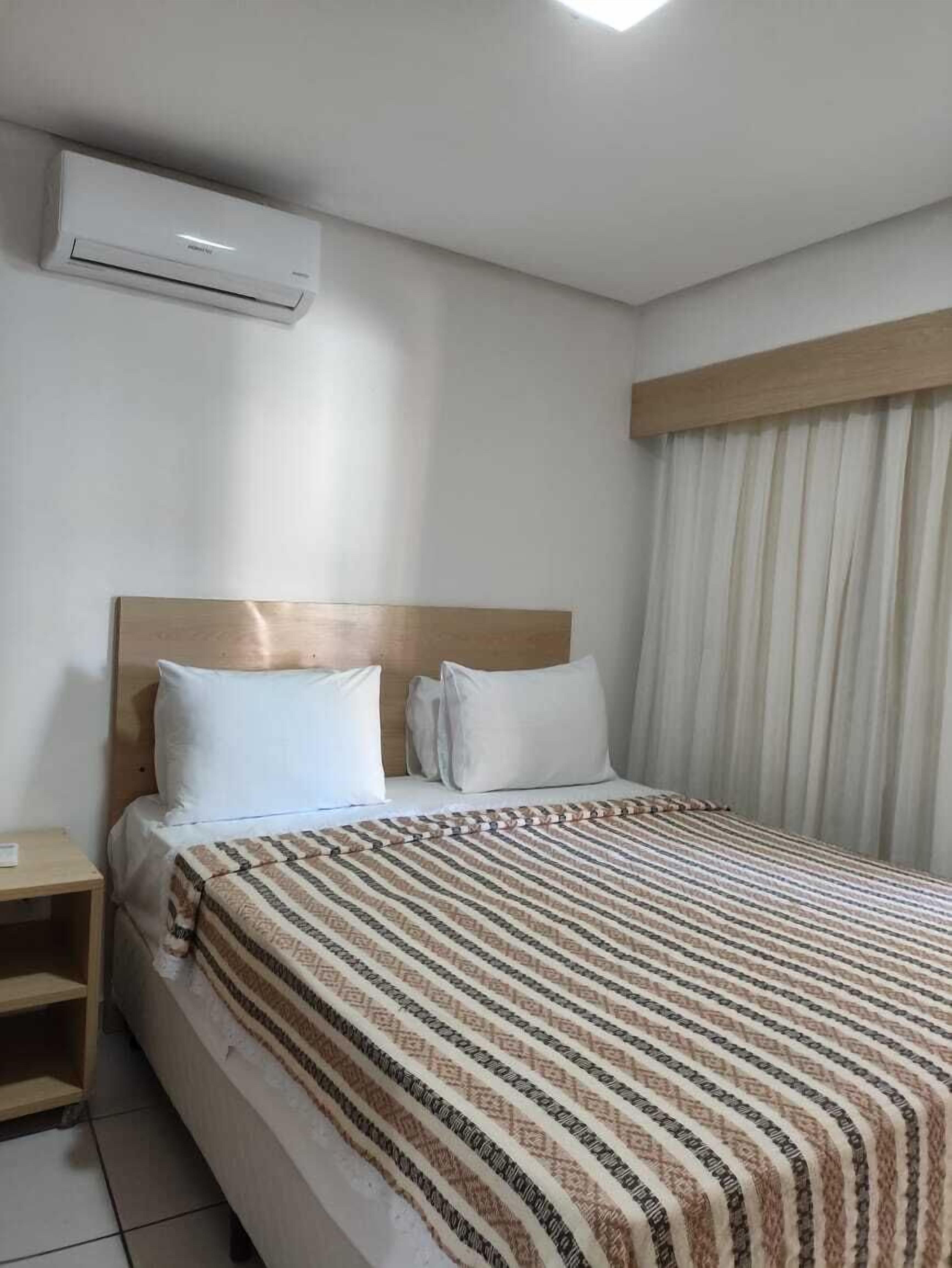 Hotellihuone, jossa on sänky, yöpöytä, puinen pääty, ikkuna verhoilla ja ilmastointilaite.