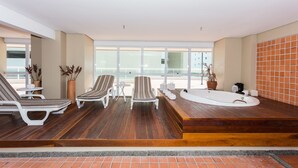 A modern indoor pool area with wooden flooring, white lounge chairs, a hot tub, and large windows offering views. - Vip Beira Mar Residence (Fortaleza)