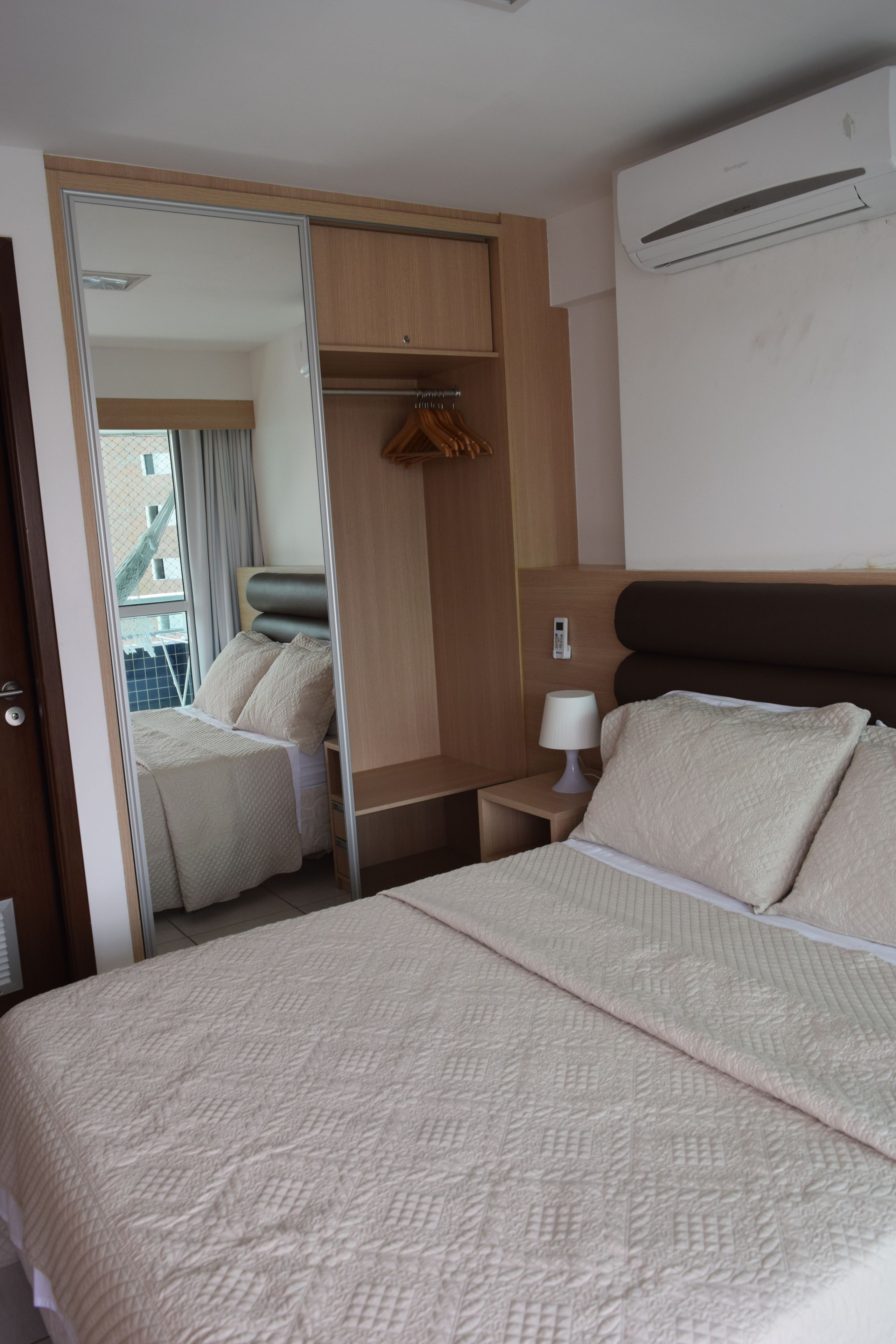 A hotel room with a bed, a wardrobe, a bedside table with a lamp, and an air conditioning unit.