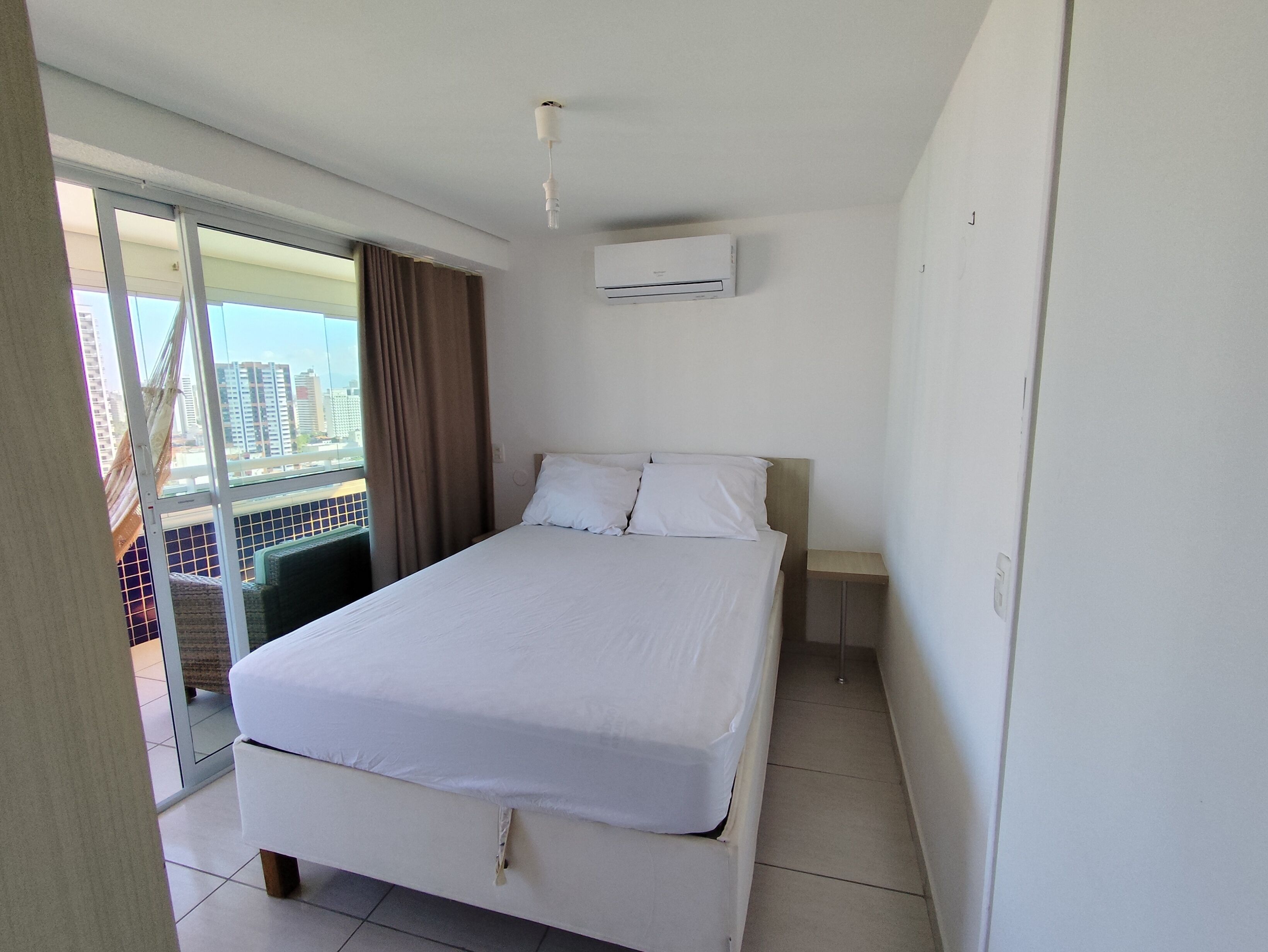 Habitación de hotel con cama, mesita de noche, equipo de aire acondicionado y vista a la ciudad a través de un ventanal grande.