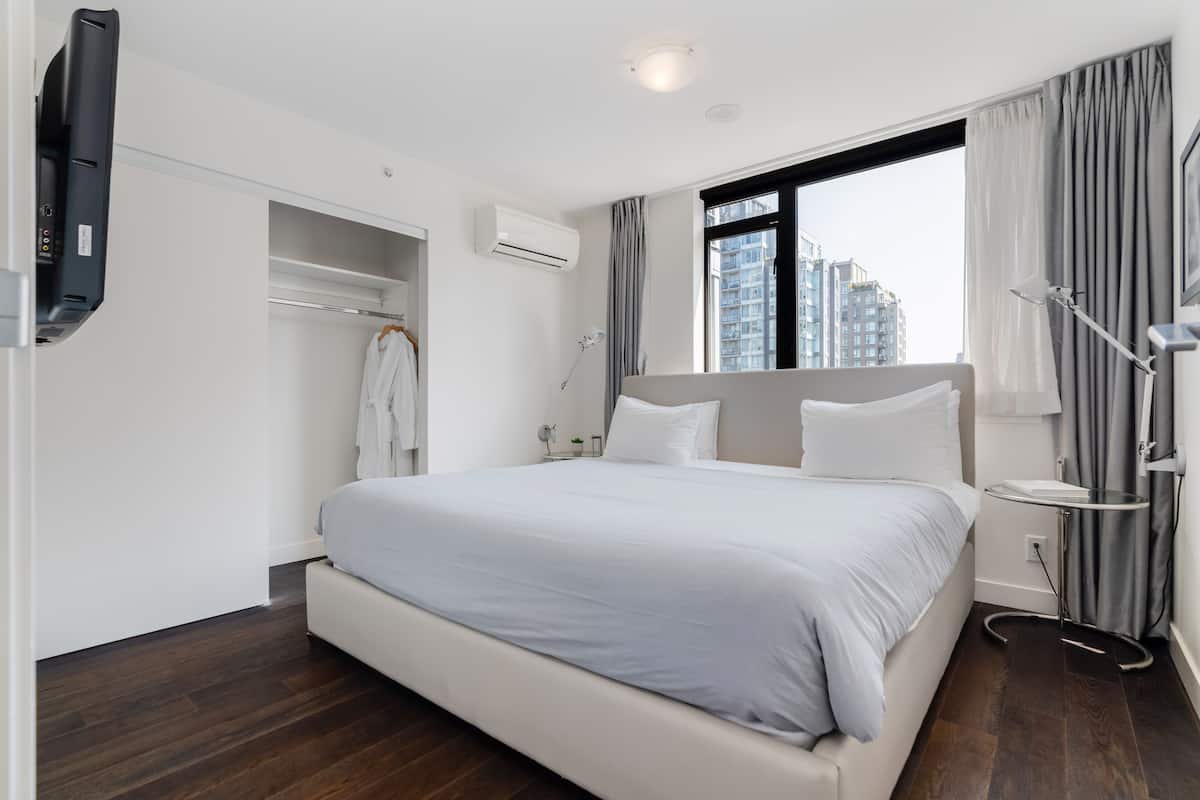 A modern hotel room with a large bed, a bedside table, a TV, and a view of the city.