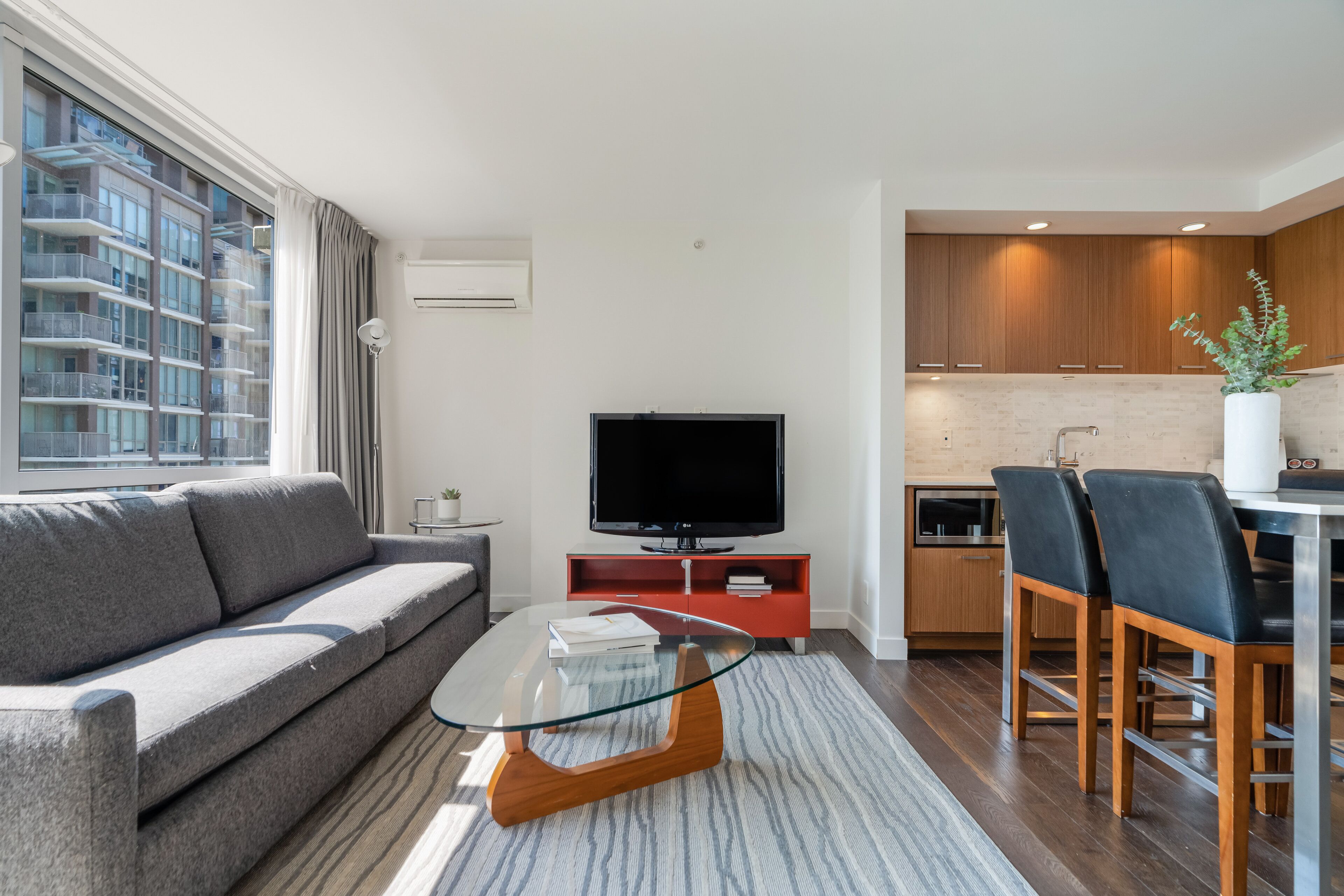 A modern living room with a sofa, a glass coffee table, a flat-screen TV, and a kitchen area with wooden cabinets and bar stools.