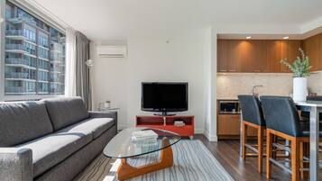 A modern living room with a sofa, a glass coffee table, a flat-screen TV, and a kitchen area with wooden cabinets and bar stools.
