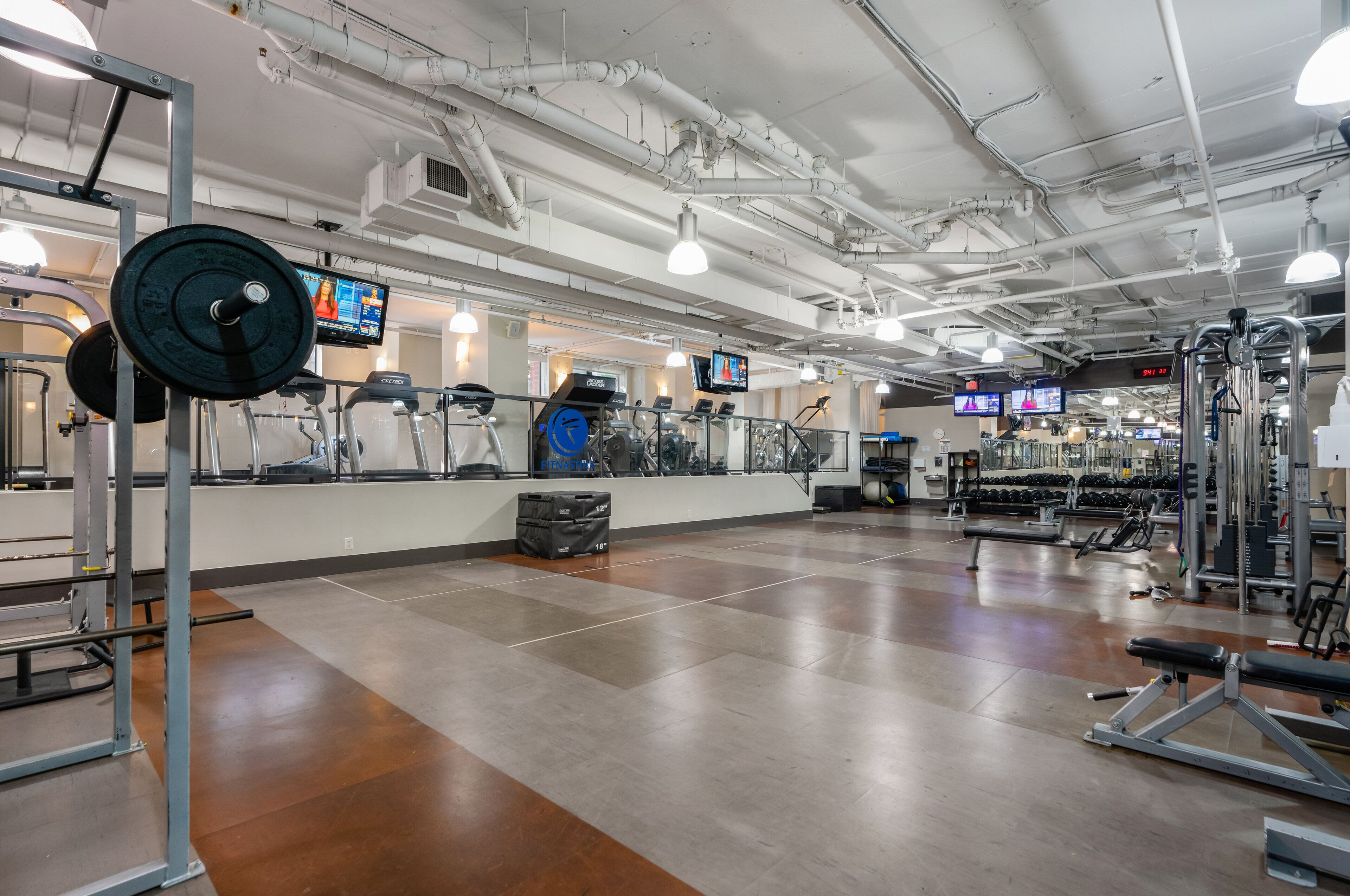 A gym with various exercise equipment, a weight rack, and multiple TV screens displaying workout videos.