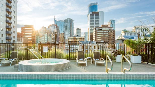 Ein Pool auf dem Dach mit Liegestühlen und einem Whirlpool, umgeben von einer Umzäunung und Stadtgebäuden im Hintergrund.