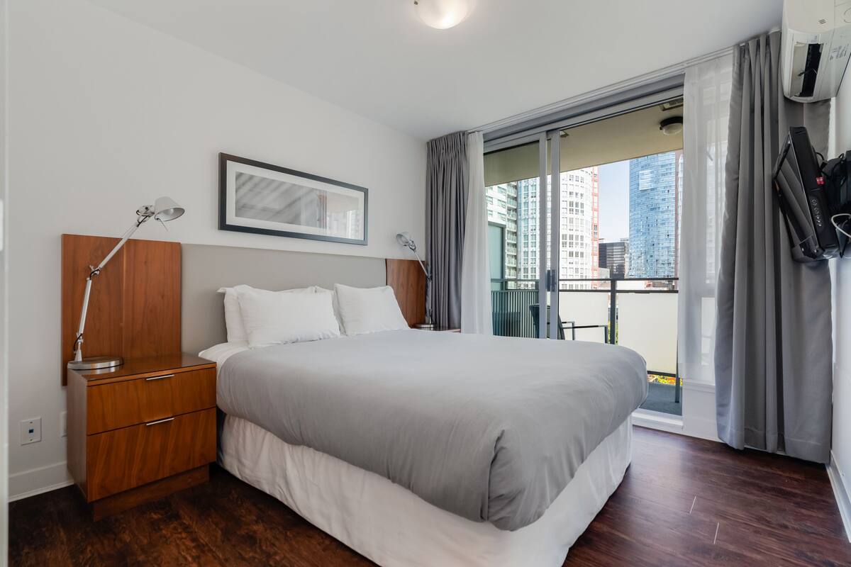 A modern hotel room with a large bed, a wooden nightstand, a framed picture on the wall, and a view of the city through the balcony.