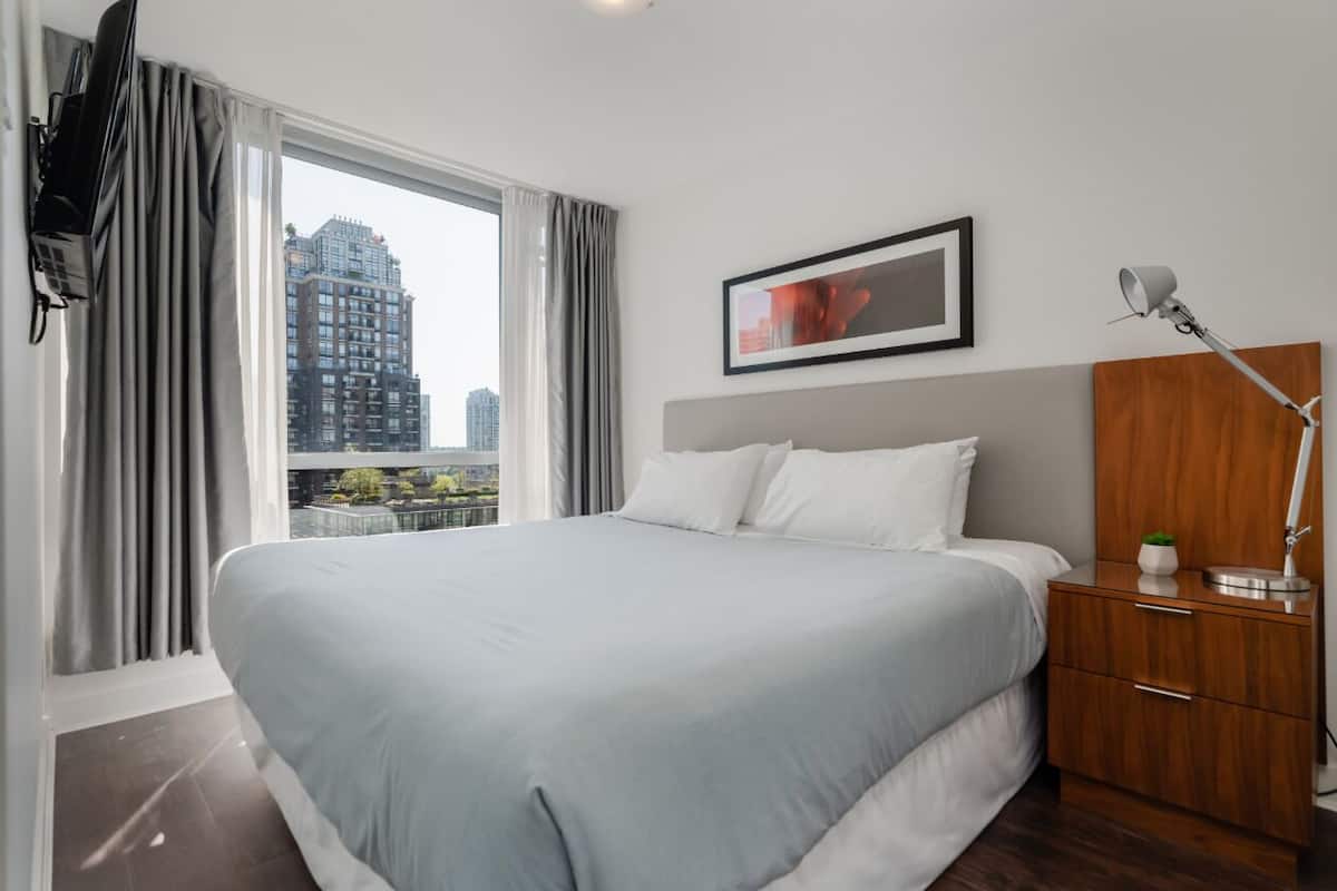 A modern hotel room with a large bed, a wooden nightstand, a desk lamp, and a city view through large windows.