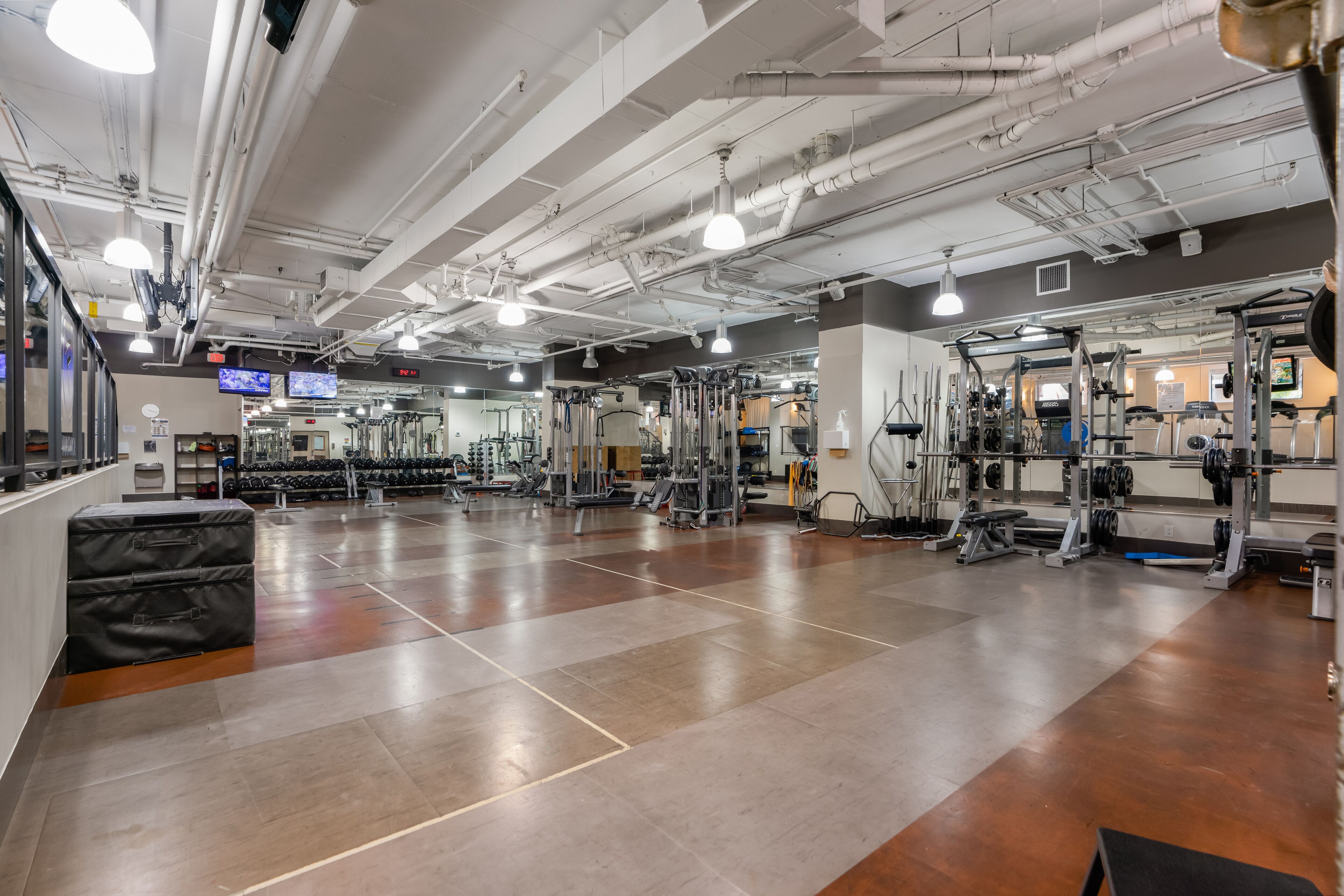 Un gimnasio espacioso con diversos equipos de ejercicio, un espejo grande y una pantalla digital en la pared.
