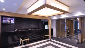 A modern reception area with a black reception desk, a digital screen, and a check-in counter. There are two wooden stools and a patterned floor. - Dormy Inn Ueno Okachimachi Hot Spring (Tokyo)