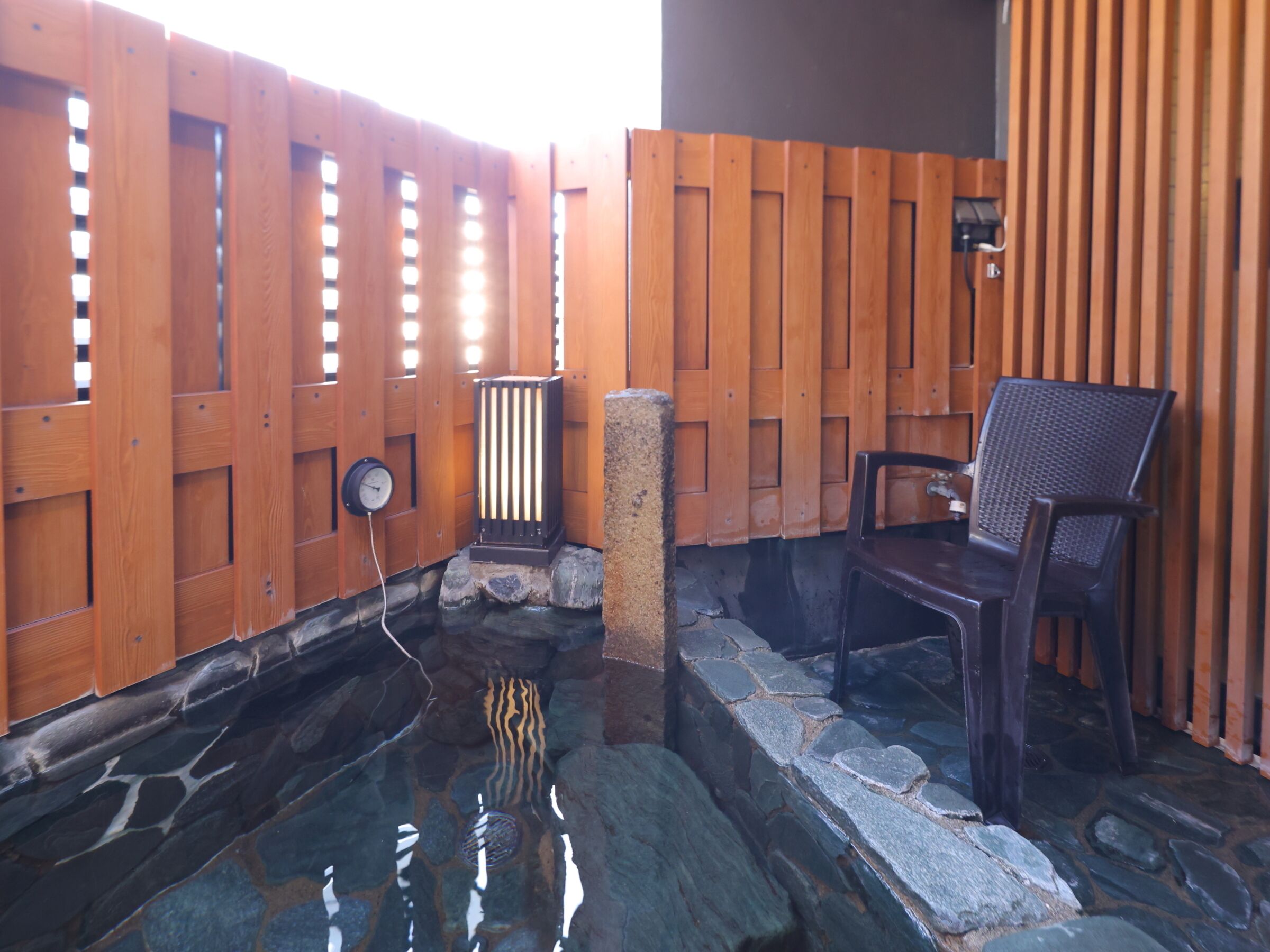 A stone hot tub area with wooden fencing, a clock, and a chair.