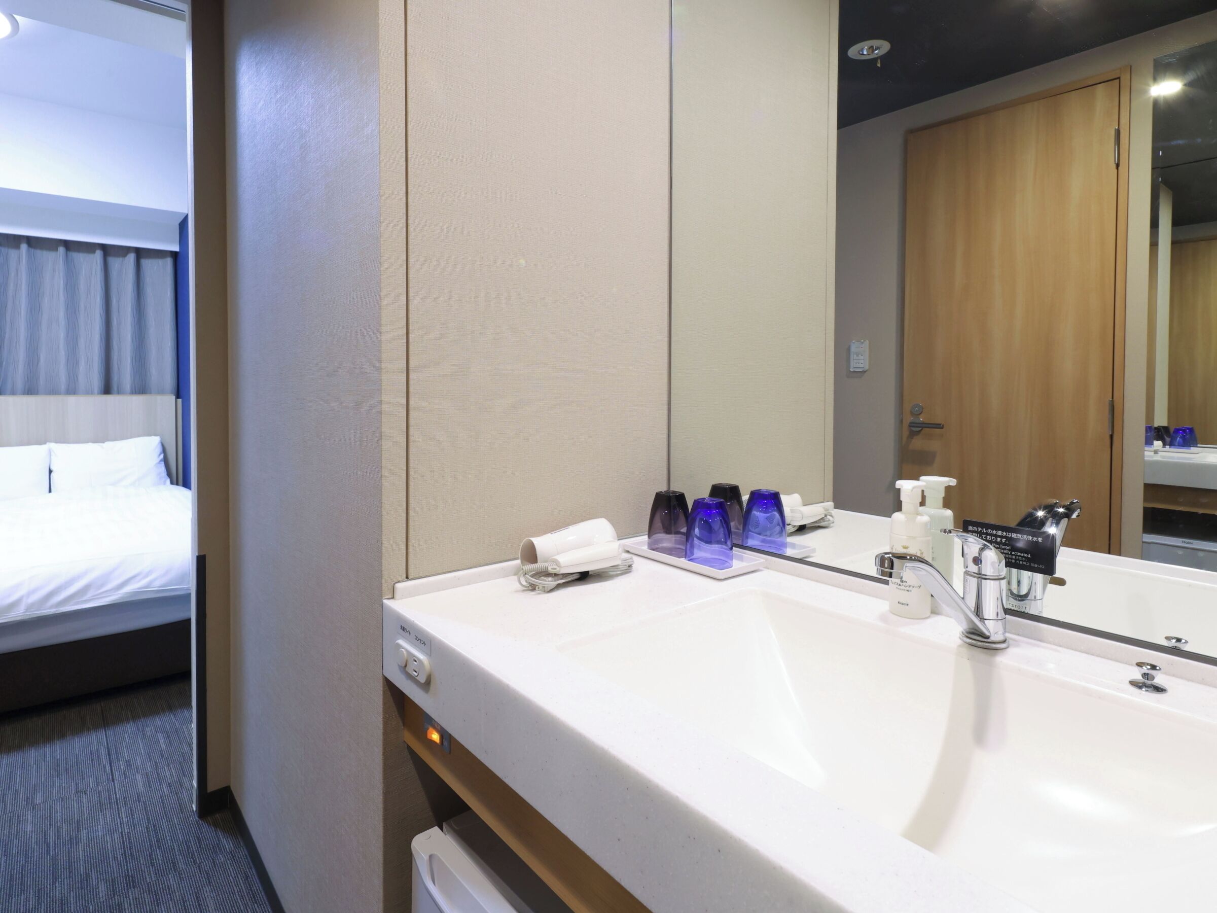 A hotel bathroom with a white sink, a mirror, and a bed with white linens.