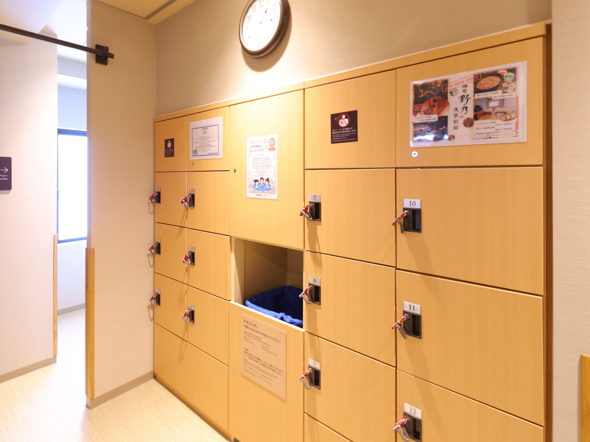 A row of wooden lockers with numbered doors, a clock on the wall, and a poster with food images.