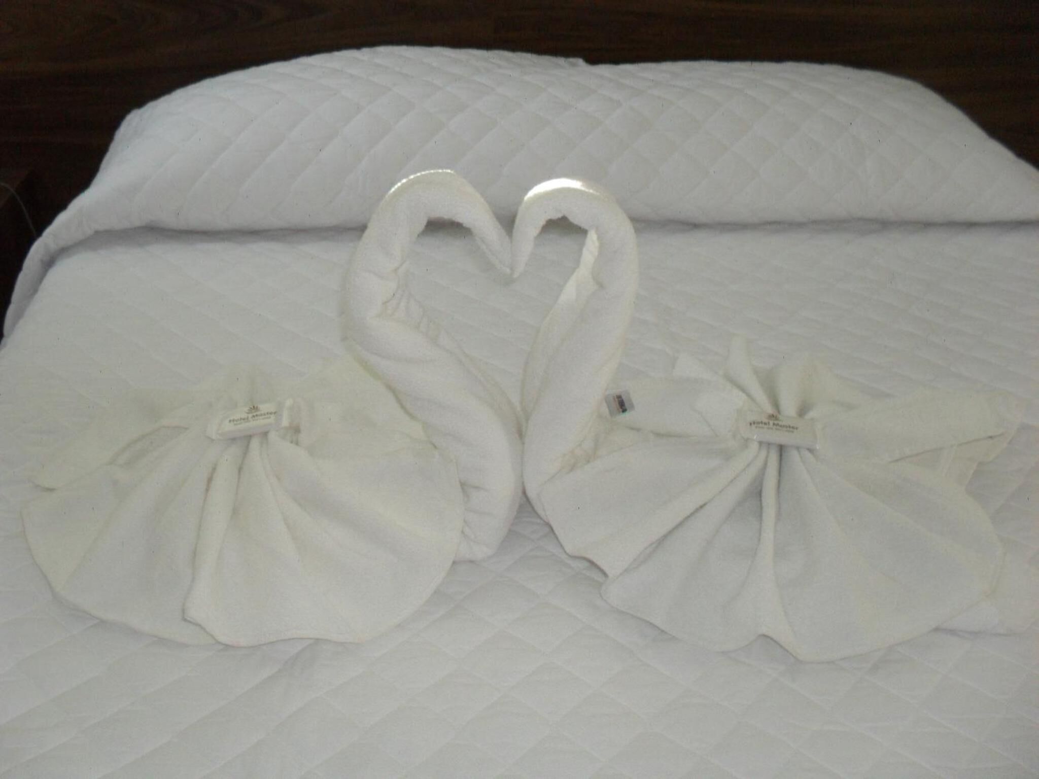 A bed with a quilted white duvet and two towels arranged in a heart shape.