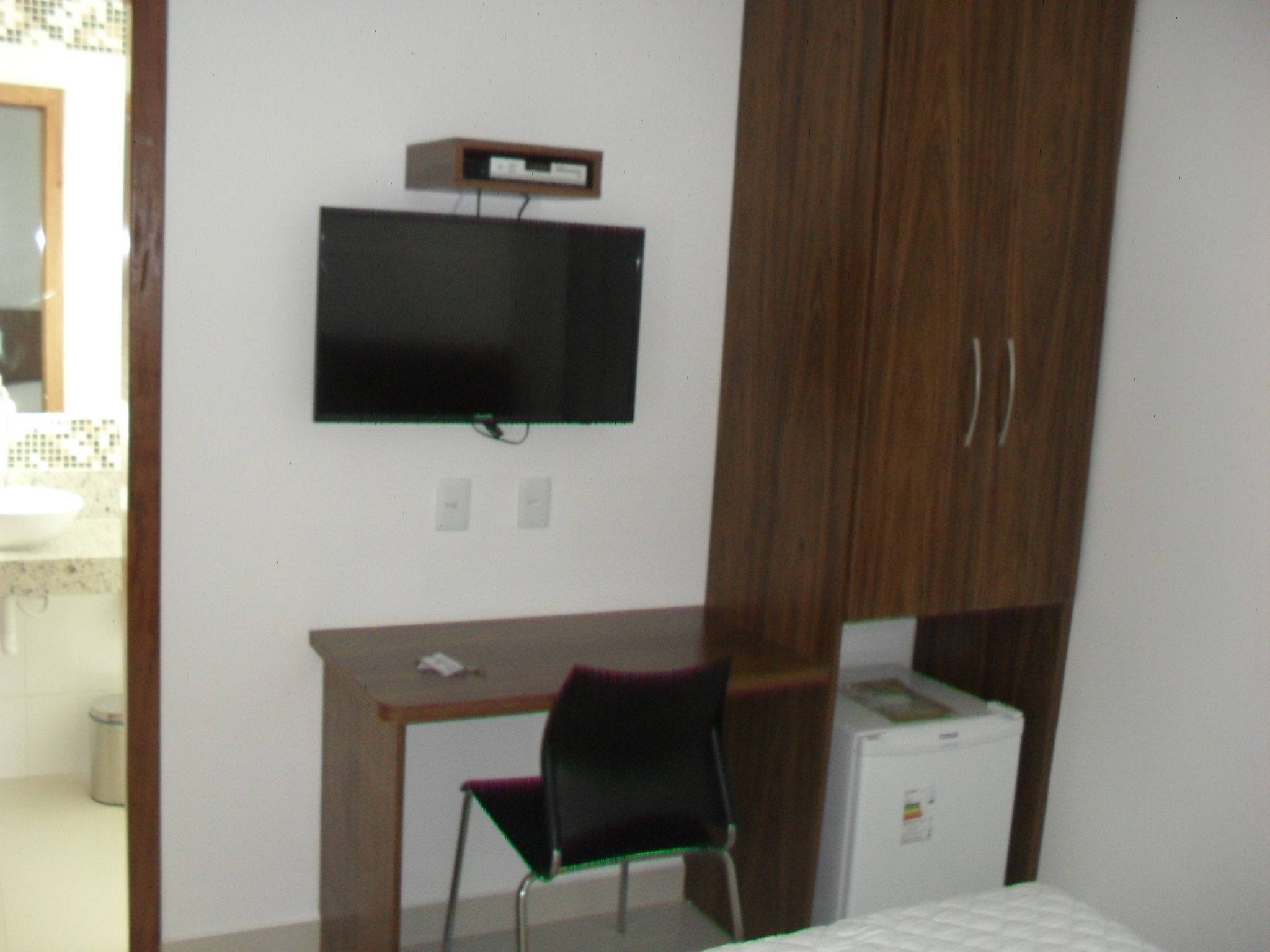 A bedroom with a bed, a desk with a chair, a television mounted on the wall, and a small refrigerator.