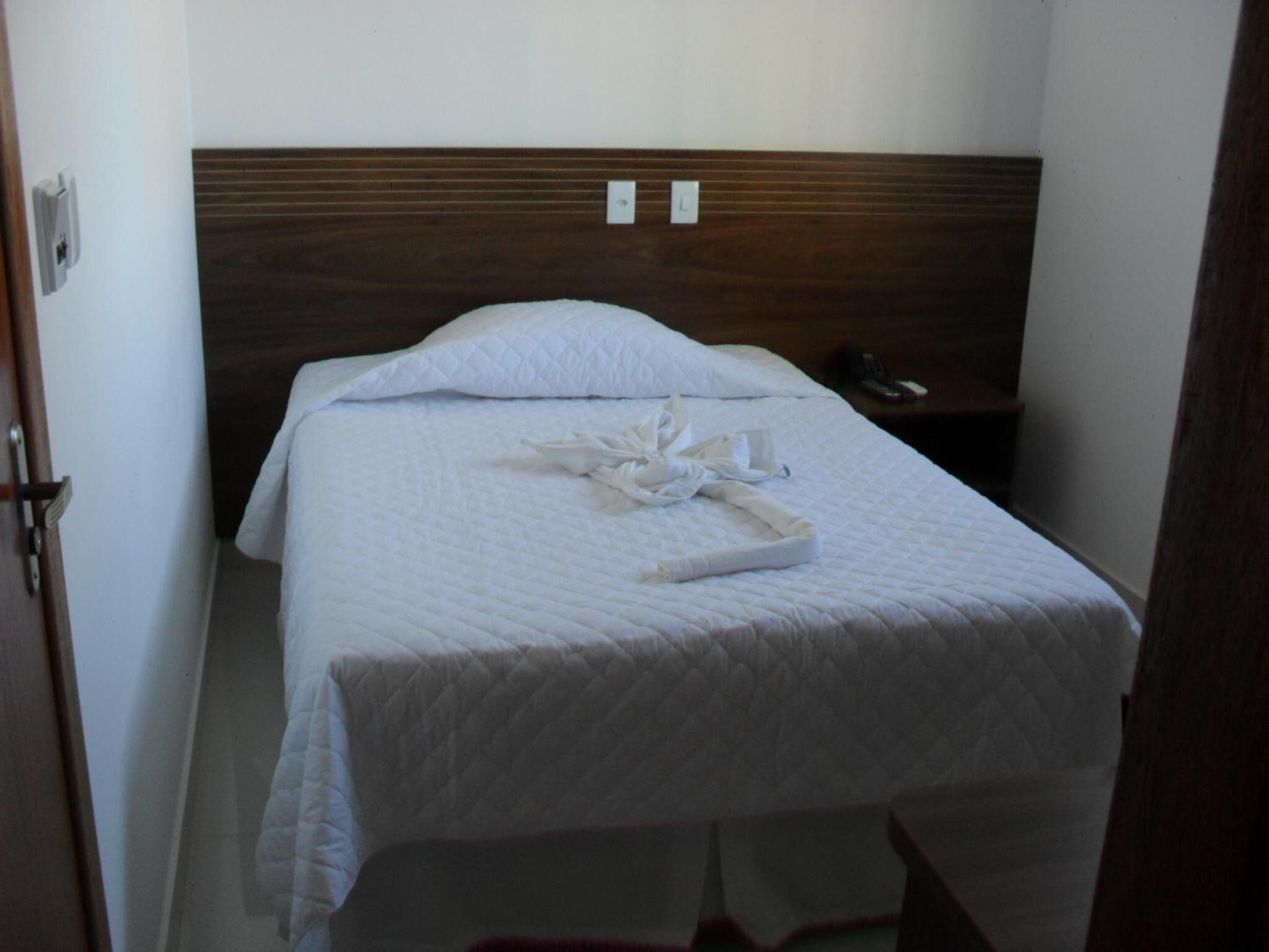 A neatly made bed with a white quilt and a towel on top, a wooden headboard, and a bedside table with a phone.