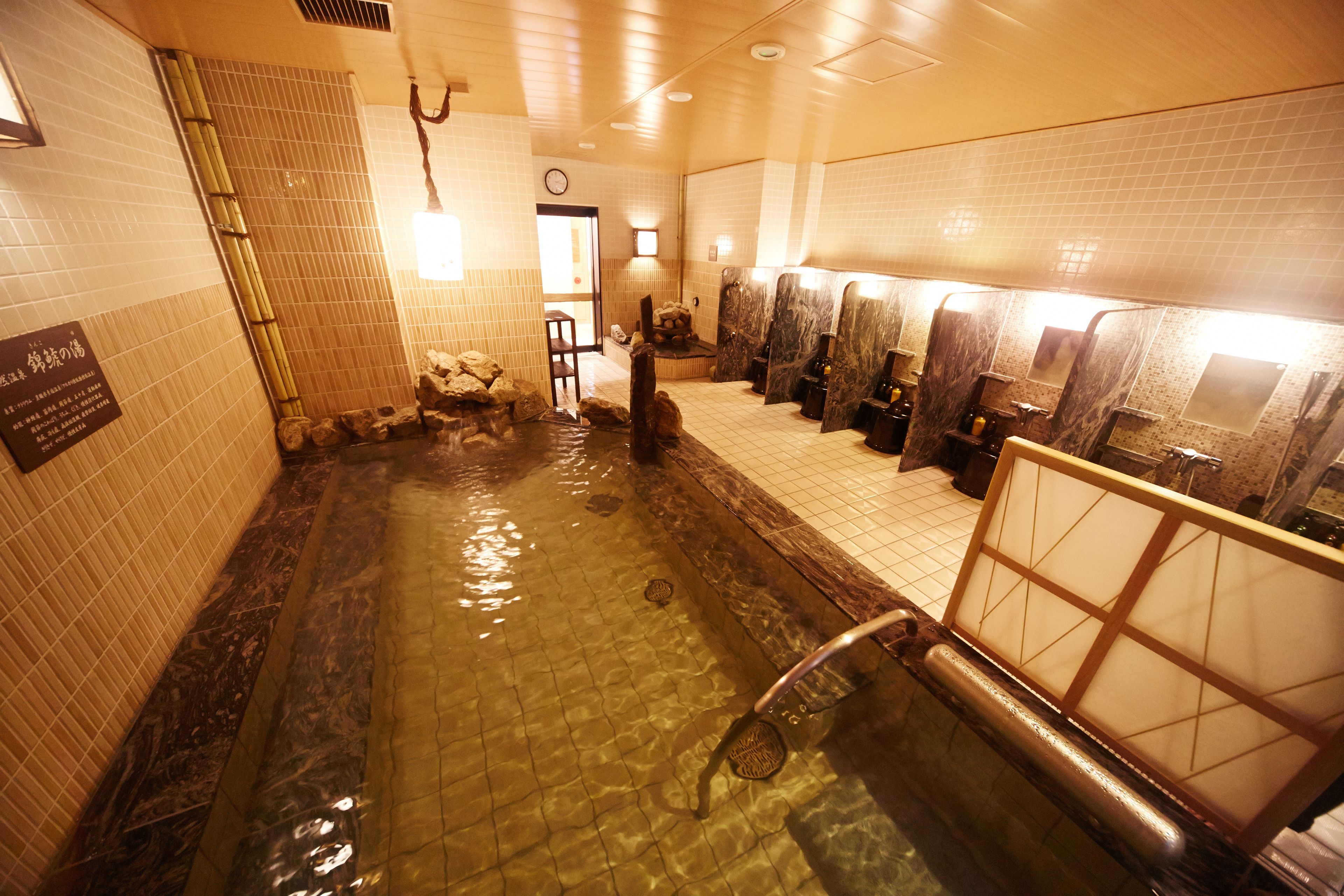 Ein öffentlicher Badebereich mit einem steingefliesten Pool, mehreren Waschbecken und einer hölzernen Bank.