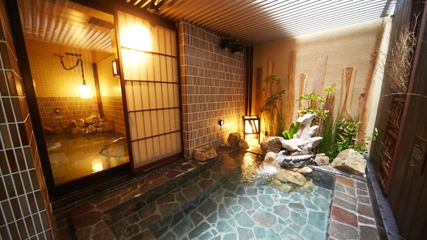 Un onsen japonais traditionnel, avec une source d’eau chaude alimentée par des pierres, des portes coulissantes en bois et un petit jardin de rocaille.
