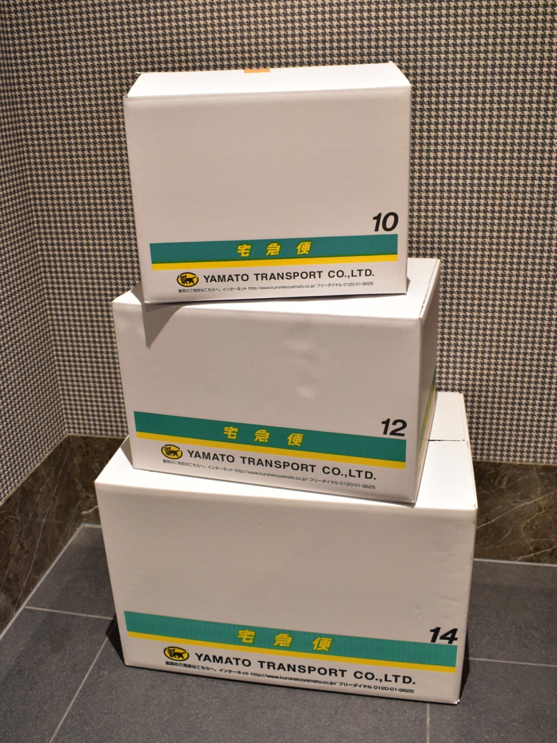 Trois boîtes empilées, portant les numéros 10, 12 et 14, et sur lesquelles figurent le texte « YAMATO TRANSPORT CO., LTD. » ainsi qu’une adresse de site web.