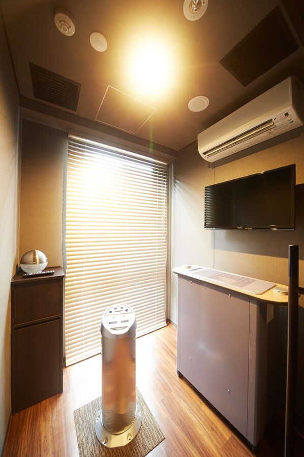 A compact hotel room with a wooden floor, a desk with a flat-screen TV, a small cabinet, a standing heater, and a window with blinds. - Dormy Inn Premium Nagoya Sakae Natural Hot Spring (Nagoya)