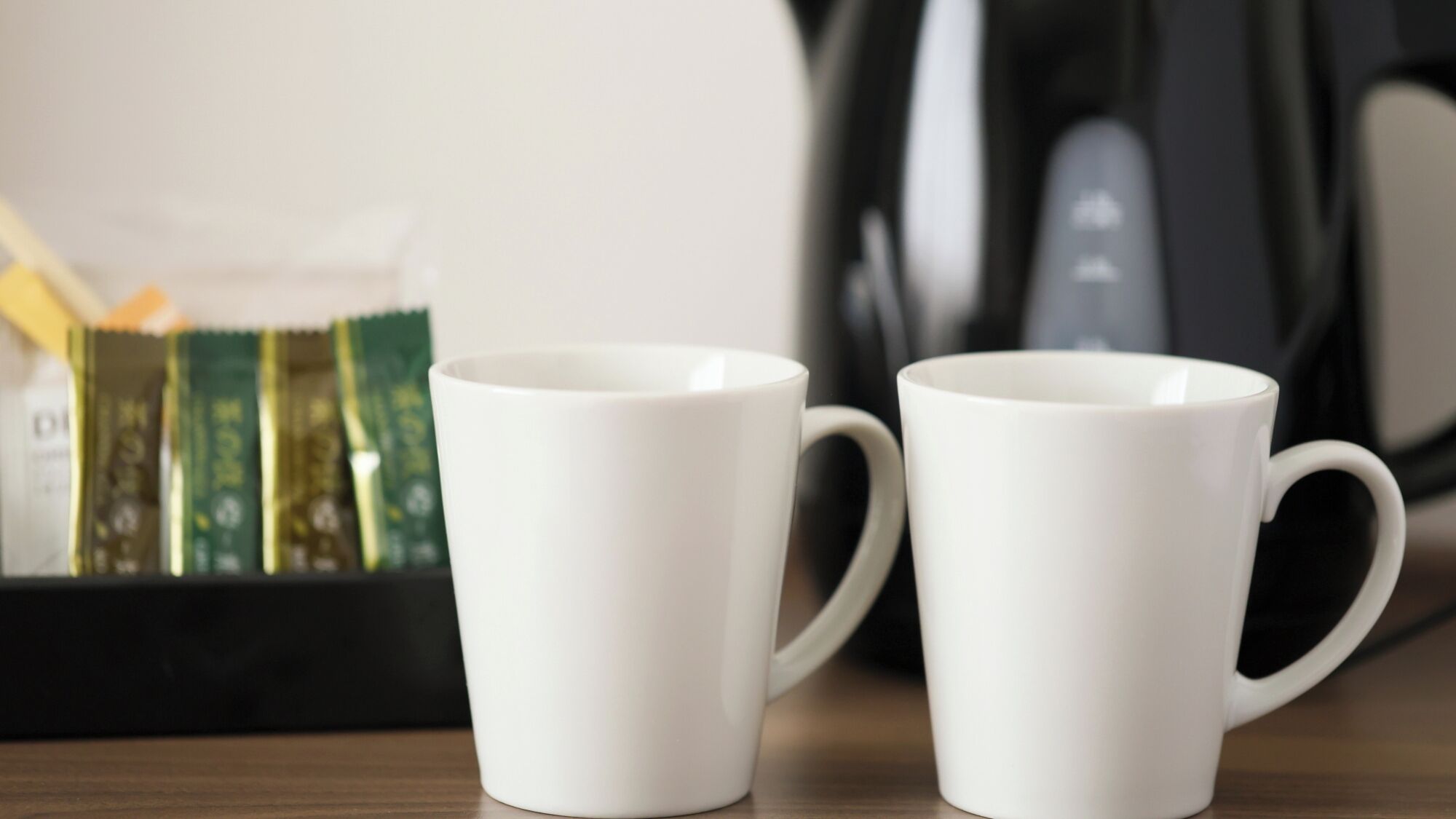 Two white ceramic mugs on a wooden surface with a blurred background of a coffee maker and a box of tea bags.