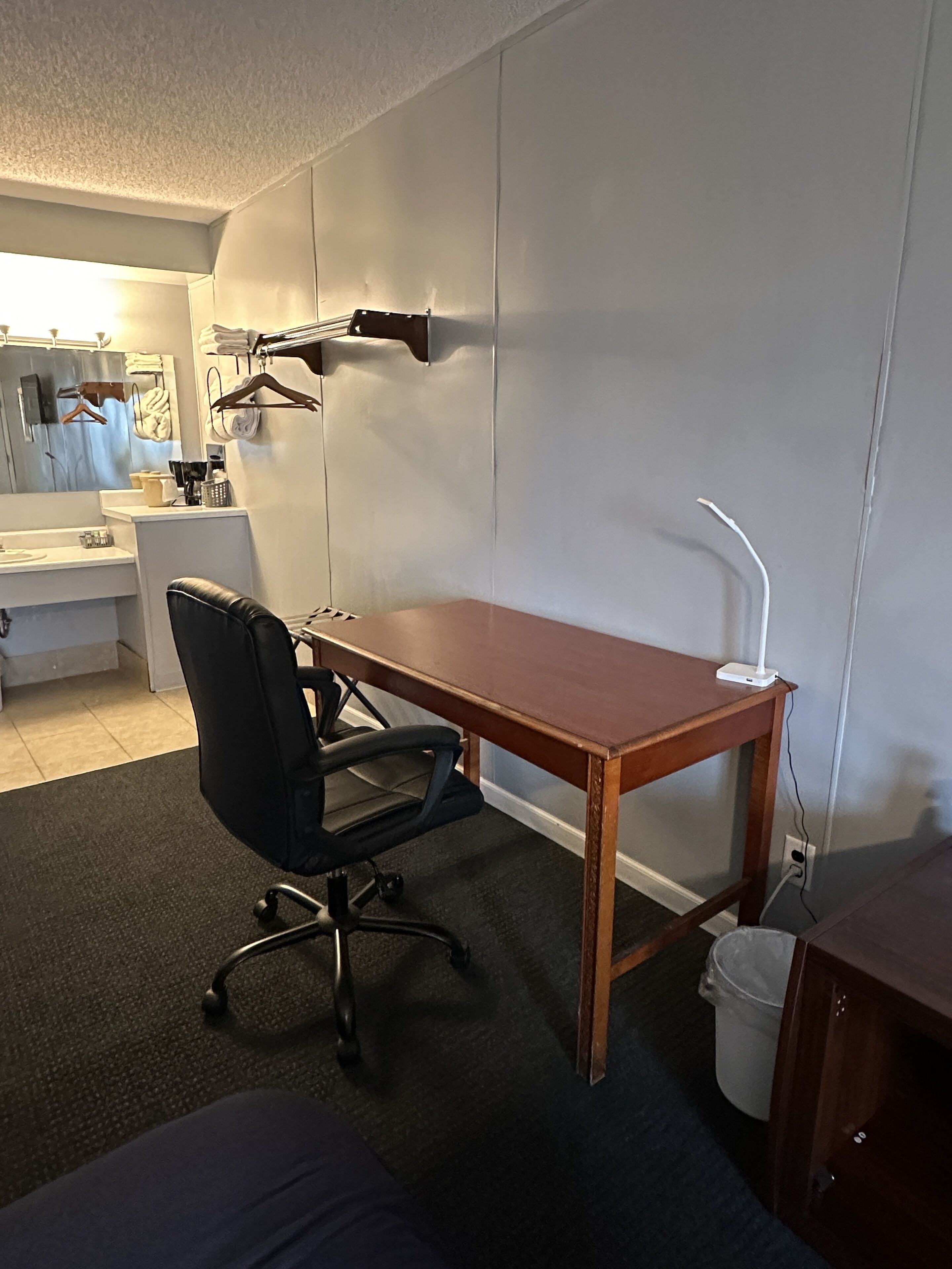 Una habitación pequeña y bien iluminada con un escritorio de madera, una silla de oficina negra, un cubo de basura blanco y un mueble pequeño.