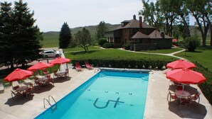 Una piscina con sombrillas y sillas rojas, rodeada de un césped cuidado y una casa grande al fondo.