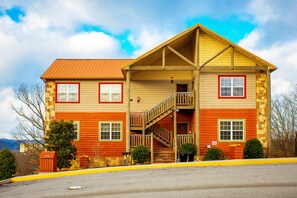 A two-story log cabin with a wooden staircase, a porch, and a mountain backdrop.